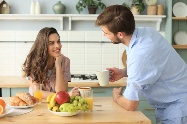 Beautiful young couple in pajamas is looking at each other and smiling while cooking in kitchen at home. - Photo, Image