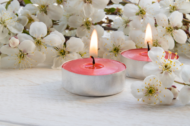 Una vela está sobre una mesa blanca de madera cerca de las ramas de flores blancas de cerezo
. - Foto, Imagen
