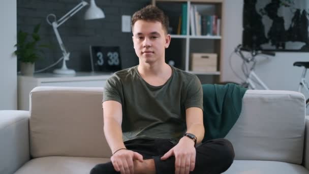A teenager is sitting on a sofa in a cosy room and turns his face to look at the camera. - Video