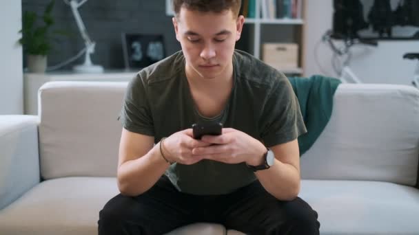 Handsome teenager is typing on his smartphone and sitting on a couch. - Materiał filmowy, wideo