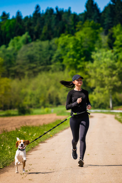 Sport girl is running with a dog (Beagle) on the rural road towadrds camera. - Φωτογραφία, εικόνα