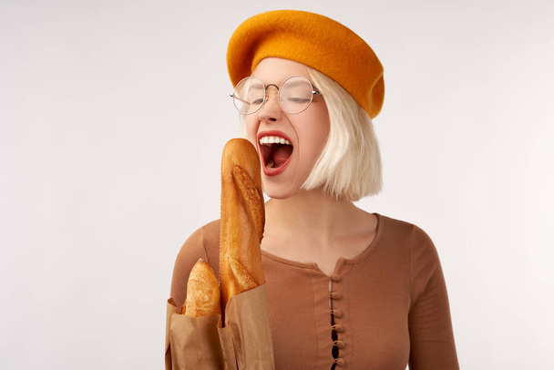 Portrait of a beautiful young woman holding baguette in her hands. Cheerful woman in yellow beret isolated over white background in Studio - Fotó, kép
