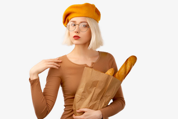 Young woman holding a bag with baguette. Serious female traveller keeps arm on shoulder, looks self assured, dressed in brown shirt, yellow beret, large round glasses, ready to explore unknown city - Fotografie, Obrázek