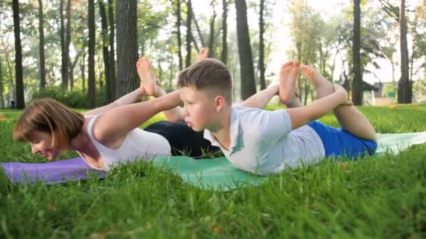 4k-beelden van tiener jongen die yoga beoefenen met middelbare leeftijd moeder op gras in Park. Familie die fitnessoefeningen doet en zich uitstrekt in het bos - Video
