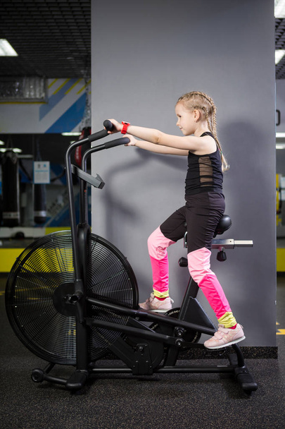Small attractive caucasian child using exercise bike in the gym. Fitness. A little athlete using an air bike for a cardio workout at the crossfit gym. Sport girl sitting on bicycle machine - Photo, Image