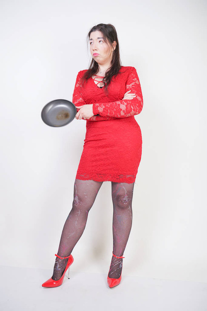 angry negative woman with a chubby body stands in a red dress and holds a black skillet on white studio background. the girl is angry and dissatisfied with the diet and her late coming husband. - Zdjęcie, obraz