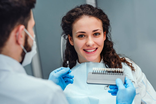 Choice of tooth color with a special scale. Dentist select a shade of tooth enamel for the young pretty girl of the patient. Bleaching, prosthetics, orthopedic dentistry. - 写真・画像