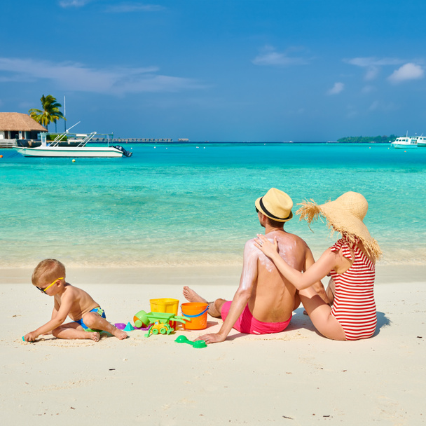 Familia en la playa, pareja joven con un niño de tres años. Mujer que aplica una loción de protección solar en la espalda del hombre. Vacaciones de verano en Maldivas. - Foto, Imagen