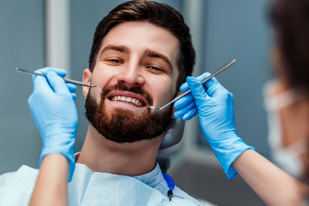 Portrait of smiling young man with dentist holding dental tools at clinic. Close up view. - 写真・画像