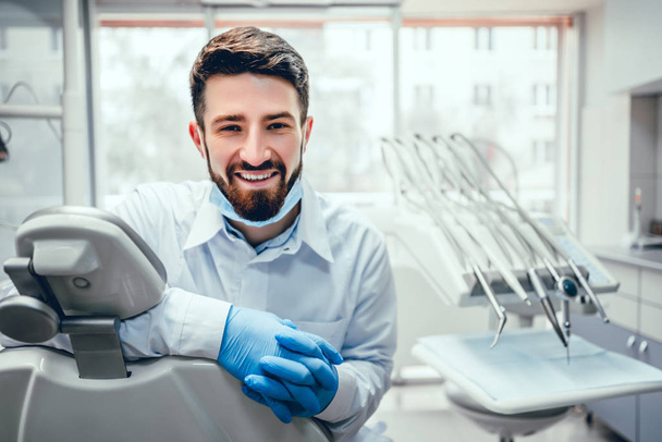 Front view of professional male dentist in white doctor coat and protective gloves sitting in dental chair and equipment, looking at camera and smiling. Bearded man posing during working process. - Фото, изображение