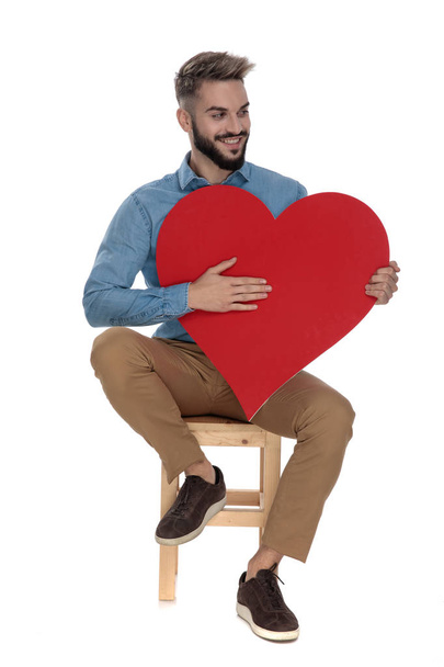сидячий привабливий чоловік тримає руку на великому червоному серці
 - Фото, зображення
