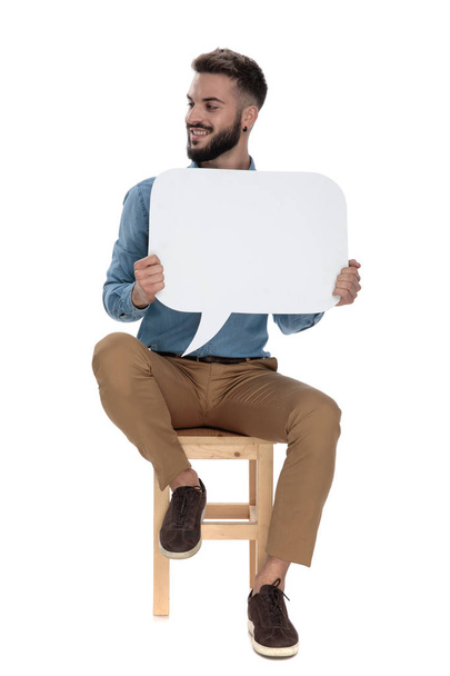 καθισμένος αλαζονικός άνθρωπος κρατώντας κενό σημάδι μήνυμα ενώ αναζητούν AWA - Φωτογραφία, εικόνα