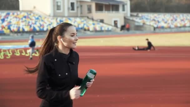 Молодая девушка бежит на беговой дорожке стадиона с бутылкой в руке
 - Кадры, видео