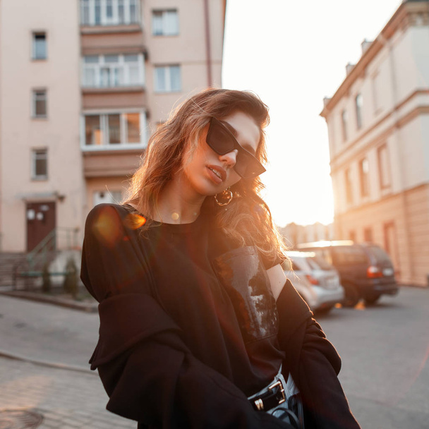 Beautiful pretty young hipster woman in trendy clothes in retro style in fashionable dark sunglasses is posing outdoors in the city on a sunny spring warm day. Urban attractive girl fashion model. - Photo, Image