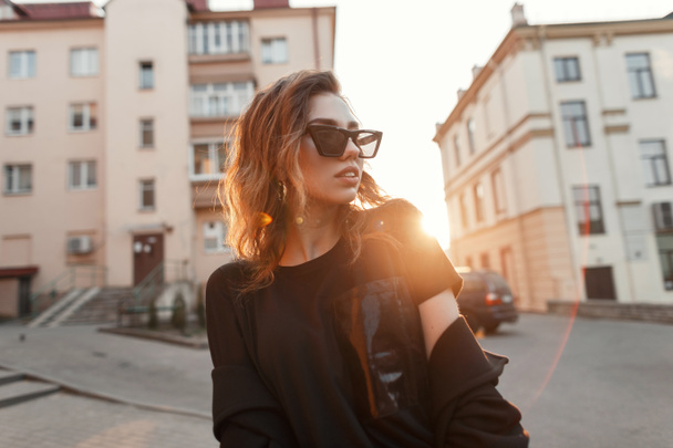 Young urban modern hipster woman in black stylish T-shirt in dark trendy sunglasses posing on the street near houses in the city. Beautiful girl model enjoys the bright orange sun. Good evening. - Foto, afbeelding