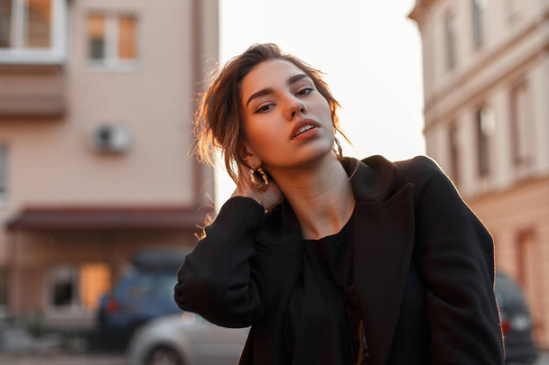 Young pretty beautiful woman in a black stylish t-shirt in a vintage coat with natural make-up with sexy lips near vintage houses in the city. Attractive girl enjoys bright orange sunshine. - 写真・画像