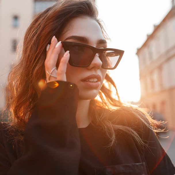 Portrait of an urban stylish woman hipster in trendy sunglasses in a stylish black coat on the background of bright orange sun highlights. Luxurious beautiful girl at sunset in the city. Close-up. - 写真・画像