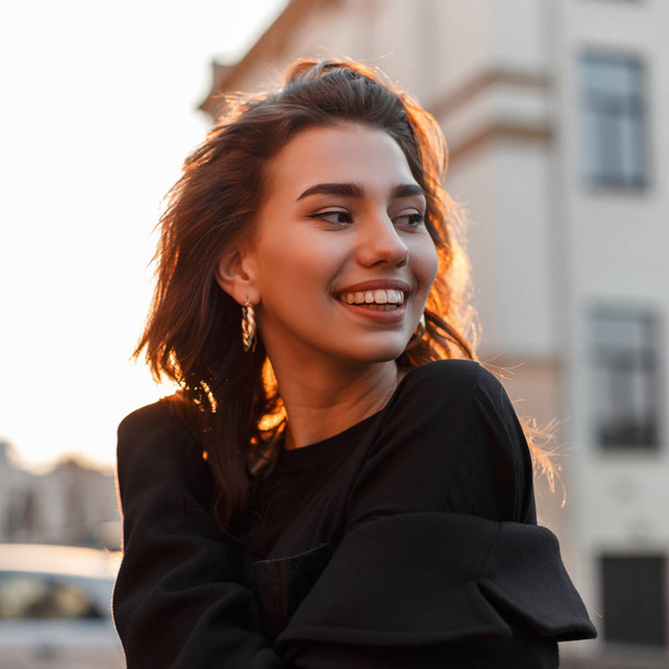 Jovencita divertida alegre en una camiseta vintage negra con pendientes dorados disfruta de una puesta de sol brillante naranja en la ciudad cerca de los edificios. Chica atractiva positiva sonríe y se relaja al sol
. - Foto, imagen