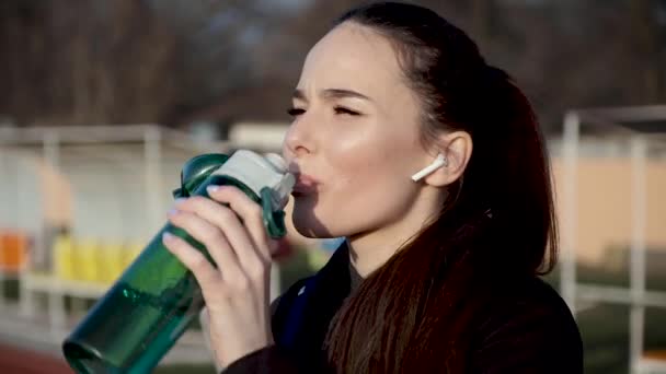 Tyttö langattomissa kuulokkeissa stadionilla juo vettä erityisestä urheilupullosta
 - Materiaali, video