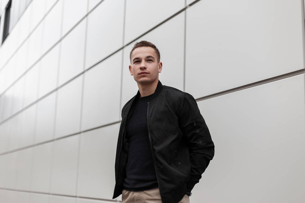 Handsome stylish young man in vintage beige pants in a black fashionable jacket in a trendy t-shirt is standing near a modern building outdoors in the spring day. European attractive guy. - Valokuva, kuva