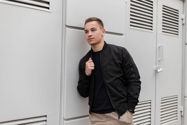 Young attractive fashionable man in a vintage stylish jacket in beige pants in a T-shirt relaxes standing near a gray metal wall near the building. Modern American is resting on a spring day. Fashion. - Fotoğraf, Görsel