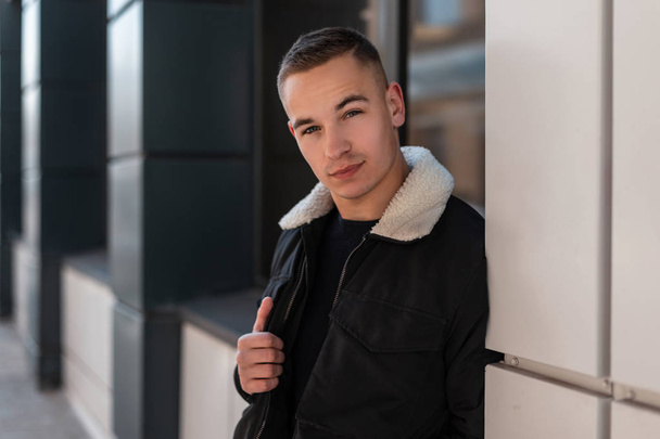 Cool young man with a trendy hairstyle in a fashionable black jacket in a T-shirt posing standing near the window of a modern building in the city. Nice guy on the street. Stylish men's clothing. - Photo, Image
