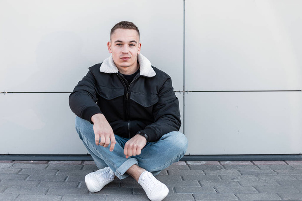 Nice young man with a stylish hairstyle in a fashionable black jacket in vintage jeans in leather trendy sneakers resting sitting on a gray tile near a modern wall on a bright spring day. Urban guy. - Photo, Image