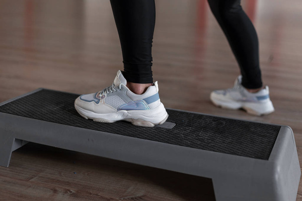 Dames benen in stijlvolle sportieve witte sneakers staan op het perron in de sportschool. Jonge vrouw traint in een fitnessstudio. Sport en gezondheid. Close-up. - Foto, afbeelding