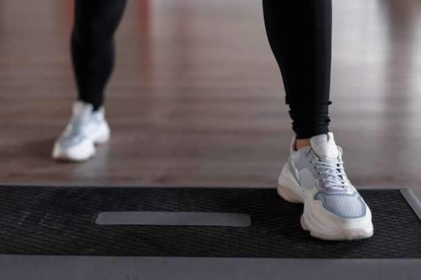 Young woman in stylish white sneakers in black leggings does exercises on the steps of the platform in the gym. Girl is engaged in aerobics. Healthy lifestyle. Close-up of female legs. - Foto, afbeelding