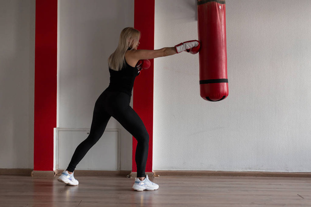 Atlética joven rubia en una camiseta negra en leggings deportivos negros en zapatillas de deporte en guantes de boxeo rojo está de pie y golpeando en un saco de boxeo en el gimnasio. Chica en entrenamiento. Estilo de vida saludable
. - Foto, imagen