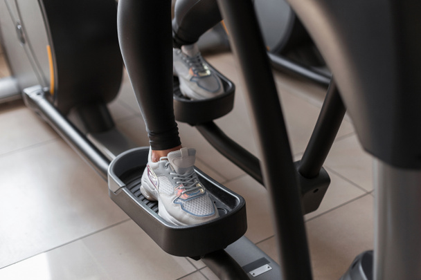 Young woman in leggings in sports sneakers does cardio training on a stepper simulator in the gym. Girl burns calories. Closeup of female legs on a modern simulator indoors. - Photo, Image