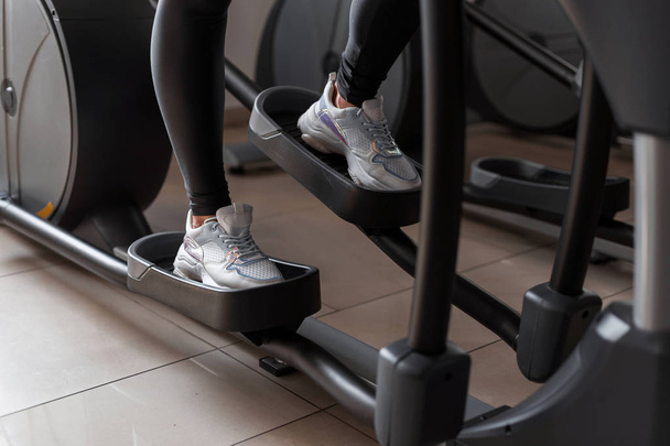 Patas de mujer en zapatillas blancas trabajando en el simulador de pasos en el gimnasio. Chica haciendo ejercicios en un club de salud. Primer plano
. - Foto, imagen