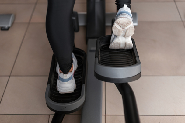 Vrouwelijke benen in witte sneakers in zwarte legging op een stepper Simulator close-up. Jonge vrouw gaat in voor sport in de sportschool. Sportief wandelen. Gezonde levensstijl. Achteraanzicht. - Foto, afbeelding