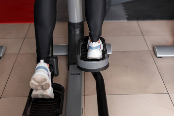 Beyaz spor ayakkabılı siyah taytlı genç bir kadın spor salonunda bir stepper simülatöründe yürüyor. Kız kalori yakmak için kardiyo yapıyor. Kadının bacaklarına yakın çekim. Arka plan. - Fotoğraf, Görsel