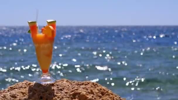 エジプトのビーチでストローとガラスのトロピカルフレッシュカクテルは、紅海の背景に岩の上に立っています - 映像、動画