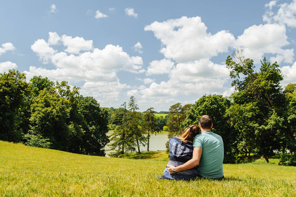 Mężczyzna i kobieta siedzący na trawie i oglądając letni krajobraz sfotografowany z tyłu - Zdjęcie, obraz