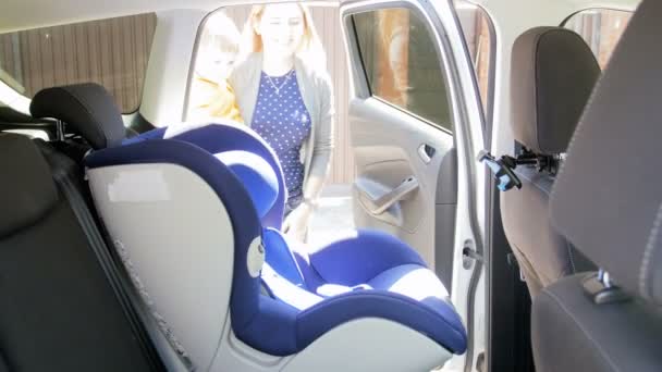 Imágenes de 4k de la madre que cuida joven que asienta a su hijo pequeño en el asiento del coche de la seguridad del niño antes de montar vehículo
 - Metraje, vídeo