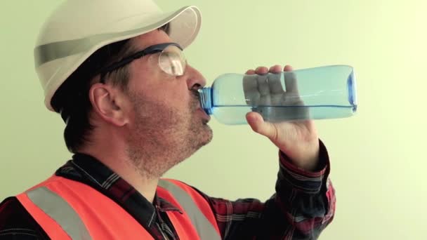 Yansıtıcı yelek, koruyucu gözlük ve beyaz kask giymiş Kafkas traşsız adam işçi, açık yeşil arka plan üzerinde izole plastik şişe su içmek ... Endüstriyel güvenlik konsepti. - Video, Çekim