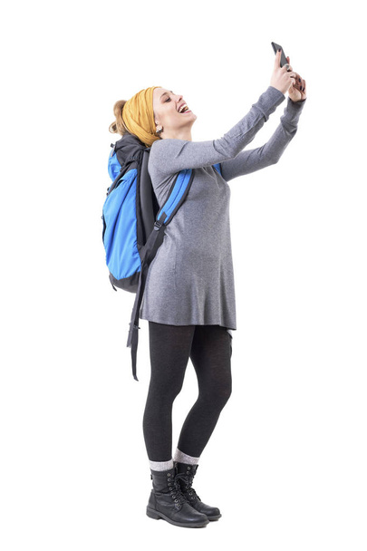 Oldalnézetből gondtalan boldog nevetés csípő turista lány hátizsák figyelembe selfies. Fehér alapon izolált teljes test.  - Fotó, kép