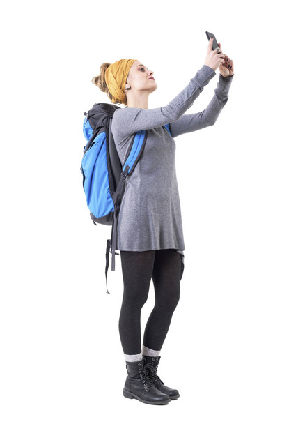 Молода хіпстерська дівчина турист з рюкзаком робить фотографії зі смартфоном, дивлячись далеко. Повне тіло ізольоване на білому тлі
.  - Фото, зображення