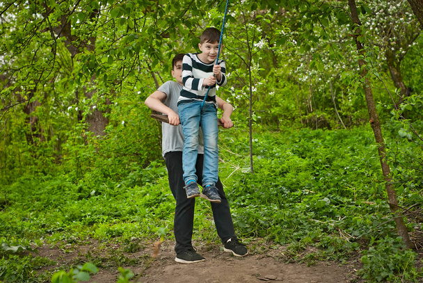Children are riding a swing in the park. Guys on a homemade swing from ropes and boards. Light blur as an artistic effect of motion. - Valokuva, kuva