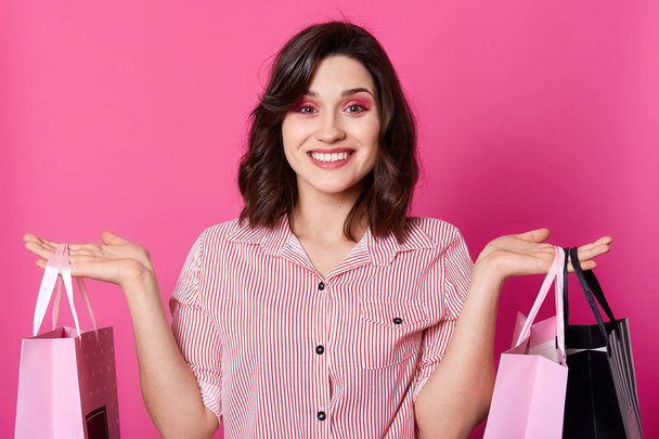 Close up portrait of happy beautiful smiling brunette woman with wavy hair, dressed striped shirt, holds shopping bags. Female model stands smiling while posing isolated over rosy studio background. - Foto, Imagen