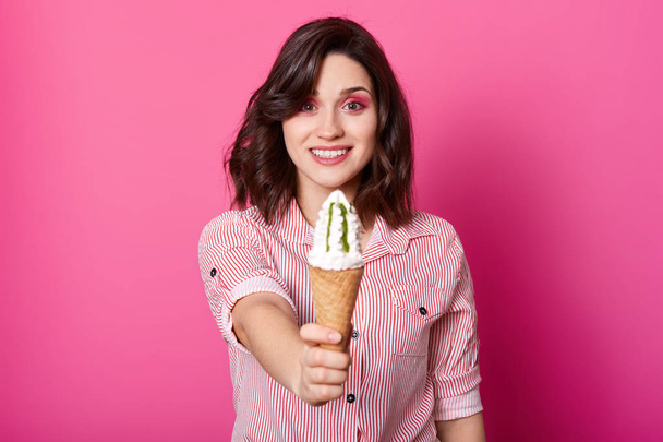 Close up portrait of pleasant looking woman offering ice cream, lady wearing striped shirt stands smiling isolated on pink background, female holding tasty cone. Copy space for advertisment. - Фото, зображення
