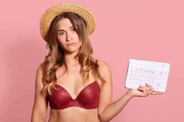Close up portrait of pleasant looking woman with upset facial expression, wears headgear and red swimsuit, holds periods calendar for checking menstruation days, poses over pink studio background. - Foto, Bild