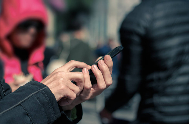 Άνθρωπος κρατώντας και πληκτρολογώντας στο κινητό τηλέφωνο στο δρόμο, πλήθος των ανθρώπων στο παρασκήνιο. Εθισμός στην τεχνολογία. - Φωτογραφία, εικόνα