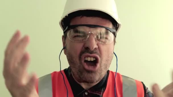 Caucasian unshaven man worker dressed in a reflective vest, protective glasses and white helmet nervously arguing, shouting, waving his arms to the camera. Isolated on light green background.. Industrial safety concept. - Footage, Video