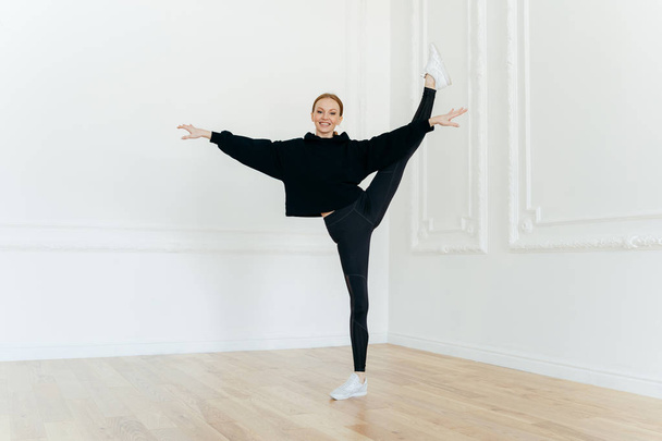 zufriedene Ballerina balanciert auf einem Bein, dehnt sich im Studio, praktiziert Yoga, zeigt gute Beweglichkeit, trägt schwarze Kleidung, hat einen glücklichen Gesichtsausdruck, führt Übungen im Innenraum aus. - Foto, Bild