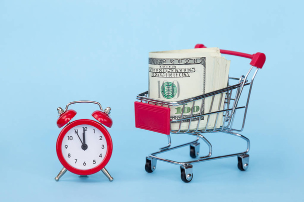 Reloj despertador rojo y un carro de supermercado con dólares sobre fondo azul
 - Foto, Imagen