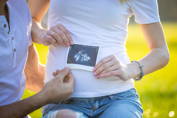 Happy couple expecting baby, pregnant young woman with husband sitting on grass and holding ultrasound pictures, young family and new life concept - Фото, изображение