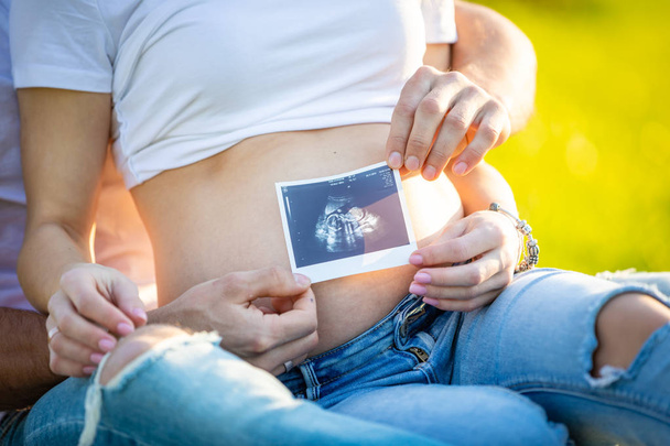 glückliches Paar erwartet Baby, schwangere junge Frau mit Mann sitzt auf Gras und hält Ultraschallbilder, junge Familie und neues Lebenskonzept - Foto, Bild